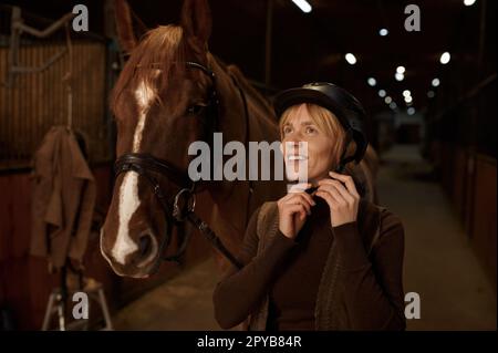 Woman rider putting helmet on head standing front of harnessed horse Stock Photo