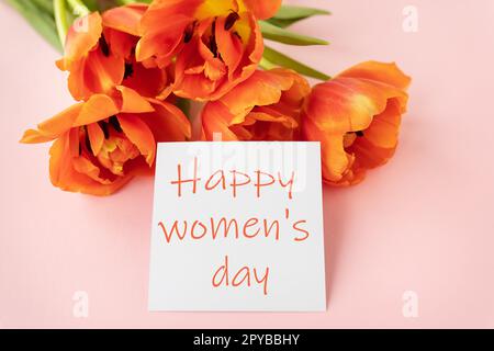 A beautiful bouquet of orange peony tulips along with a white card in the middle with the words happy women's day on a pink background. Stock Photo