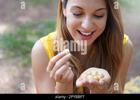 Beautiful girl eating macadamia nuts outdoor. Looks at macadamia nuts in her hand. Stock Photo