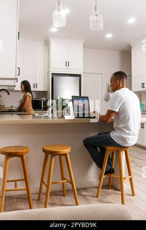 Young man working on his computer having a cup of a drink, in the background his partner doing the cleaning at the sink. Stock Photo