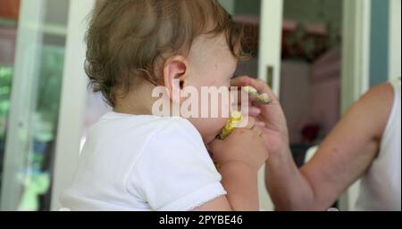 Parent feeding adorable baby infant on highchair. Cute toddler holding and eating corn Stock Photo