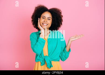 Friendly pretty, trendy curly haired african american or brazilian girl, shows her hand to the empty space for your advertisement, announcement, presentation, stand on isolated pink background, smiles Stock Photo