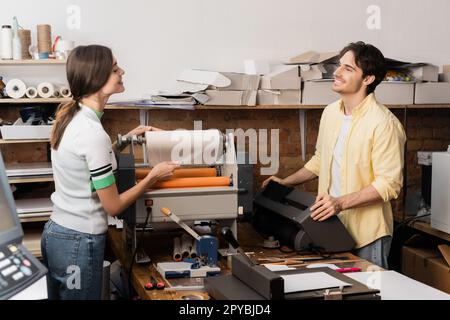 cheerful woman pulling paper while working with print plotter next to happy colleague,stock image Stock Photo