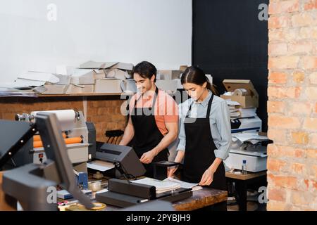 cheerful man in apron working with printer next to pretty colleague in print center,stock image Stock Photo