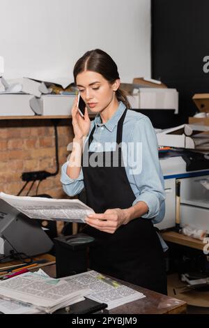 typographer in apron looking at newspaper while having phone call in print center,stock image Stock Photo