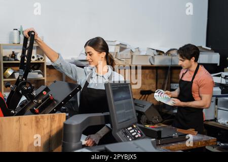 cheerful typographer in apron using professional print plotter while working with colleague in print center,stock image Stock Photo