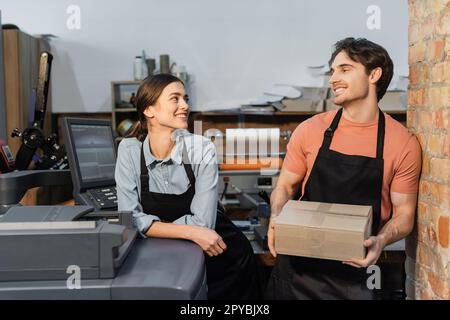 happy woman in apron leaning on printer and looking at colleague with carton box in print center,stock image Stock Photo