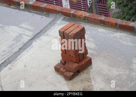 Red clay bricks or burnt clay red bricks kept in a small pile on the construction site with a view of brick arrangement in the background Stock Photo