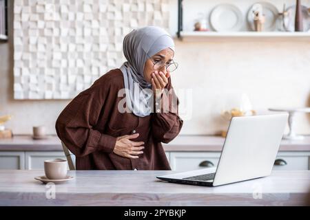 Muslim woman in hijab sitting at home in the kitchen and using a laptop. She holds her stomach with her hand, bent over from pain, covers her mouth with her hand, vomits, feels nausea, poisoning. Stock Photo