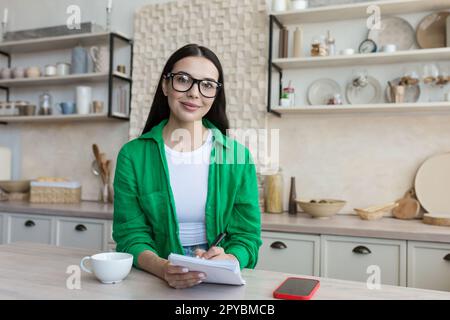 A young woman sits at home in the kitchen and writes with a pen in a notebook, keeps a personal diary, writes down dreams, makes a plan, works on self Stock Photo
