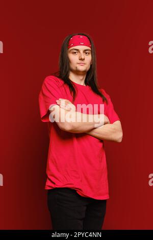 A young adult man stands in a studio with arms crossed and looks confidently at the camera against red background Stock Photo