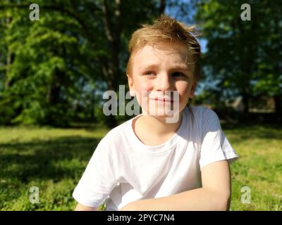 A cute young boy smiling and looks to the side. The child is dressed in a white T-shirt and blue pants. Blond hair is beautiful fall on the forehead. Long blonde eyelashes. Park in the background Stock Photo