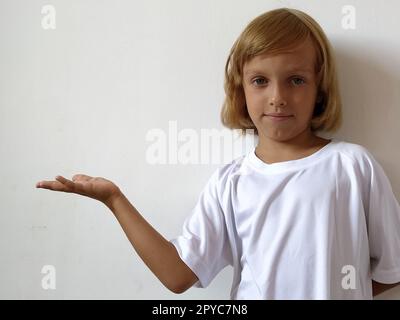 A little cute girl 6-7 years old with blond hair expresses emotional joy and a smile on a white background. A child with tanned skin. White T-shirt. Copy space. Girl with her hand points to something Stock Photo