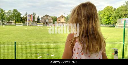 Sremska Mitrovica, Serbia, August 1, 2020. A girl sits with her back to the camera and watches a football match of childrens teams. Beautiful long blonde hair. Soccer fans in a match. Stock Photo