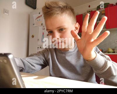 Inside shot of a blond child sitting at a table looking at the screen of a laptop or smartphone. Talk with mom or grandmother through a gadget. Online communication during self-isolation. Child waving Stock Photo