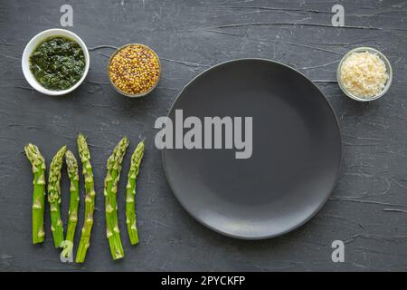Fresh asparagus with herbs and cheese and empty plate on black marble background. Stock Photo