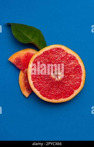 Sliced sides of a grapefruit, three pieces with a green leaf, red pulp isolated on a blue background, vertical shot Stock Photo