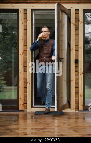 Middle age man comes out on the terrace of a small wooden house with a glass in hand Stock Photo