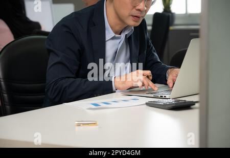Businessman working on laptop computer sitting at office desk in her workstation with colleagues in background. Stock Photo