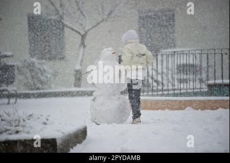 A child stands amidst a snowy scene, constructing a snowman during a blizzard. Sardinia, Italy Stock Photo