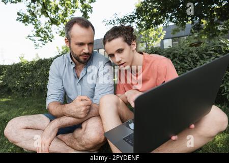 father and son looking at laptop while sitting together on green lawn,stock image Stock Photo
