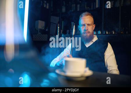 Handsome bearded man smoking hookah while drinking coffee in bar Stock Photo