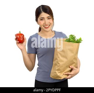 Young asian woman in casual clothes stand smiling, hold a red bell pepper while the other hand holding a paper bag full of vegetables. Portrait on white background with studio light. Isolated Stock Photo