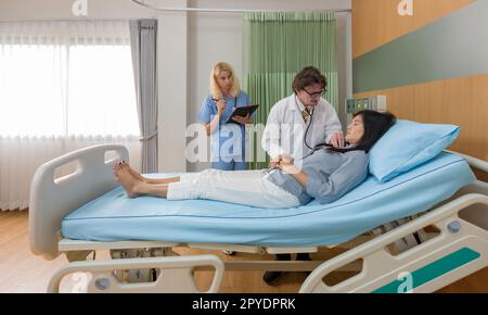 Caucasian doctor holding stethoscope checking heartbeat, examining illness of asian woman patient on the bed in hospital. Young nurse stand next to the bed, holding document clipboard. Stock Photo