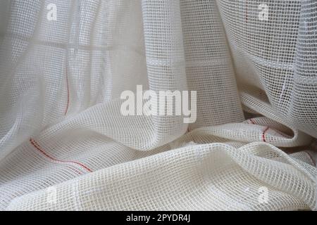 White coarse cotton or linen curtains made of weaves of thin threads. Simple compact economical apartment interior. Window decoration in the room. Red stripe. Stock Photo