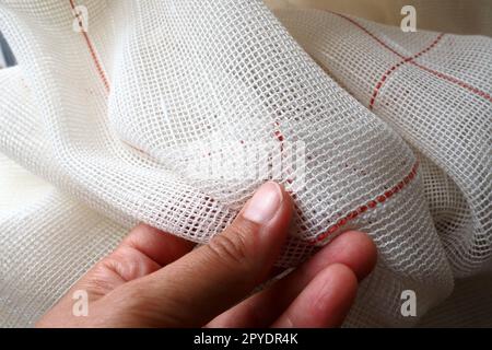 White coarse cotton or linen curtains made of weaves of thin threads. A woman's hand is feeling the fabric. Window decoration room. Red stripe. The fabric is folded in soft waves. Apartment interior. Stock Photo