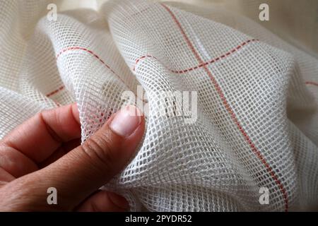 White coarse cotton or linen curtains made of weaves of thin threads. A woman's hand is feeling the fabric. Window decoration room. Red stripe. The fabric is folded in soft waves. Apartment interior. Stock Photo
