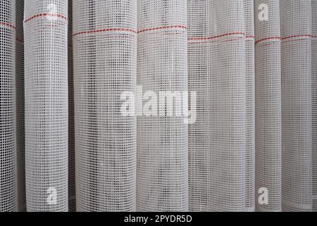 White coarse cotton or linen curtains made of weaves of thin threads. Simple compact economical apartment interior. Window decoration in the room. Red stripe. Stock Photo
