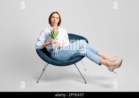 Portrait of cheerful meddle aged woman in armchair with tulips in hand Stock Photo