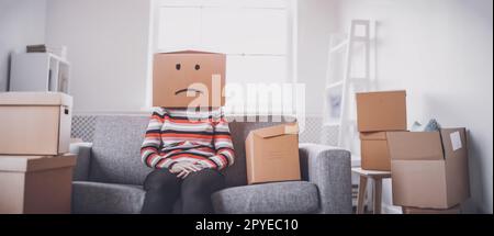 Tired sad woman with cardboard box on her head sitting on the sofa in her new house. Stock Photo