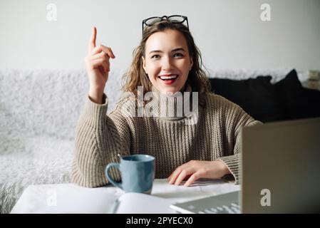 Eureka Excited female entrepreneur with successful business idea, sitting at kitchen table with laptop and showing index finger, ready to make a Stock Photo
