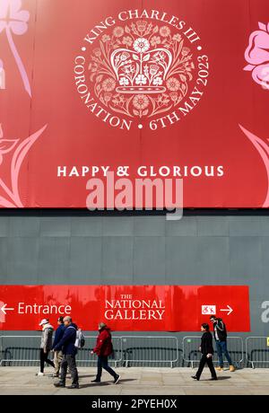 Tourists by a large red banner on the National Gallery wall with the King Charles the Third logo emblem for the coronation, Trafalgar Sq, London Stock Photo