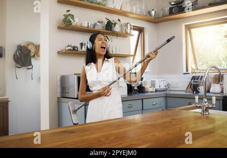 Cleaning, headphones and black woman singing and playing air guitar music with a broom in a house. Fun, playful and happy female enjoying a clean home, kitchen or room while streaming song or radio Stock Photo