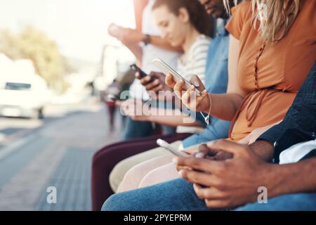 Their devices are their traveling buddies. Closeup shot of a group of people using their cellphones while sitting at a bus stop in the city. Stock Photo