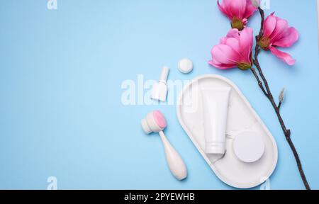 White plastic tubes and jars of cream, and a massage brush for facial cleansing on a blue background, items for cosmetic procedures Stock Photo