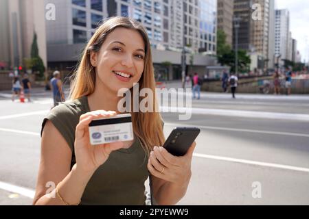 Portrait of smiling woman holding a smartphone and showing a credit card looking at camera on Paulista Avenue financial center of Sao Paulo City, Brazil Stock Photo