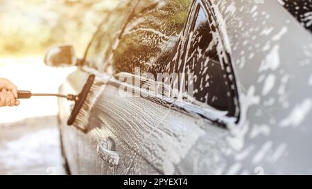 Side of silver car washed in self serve carwash, brush held by man's hand leaving strokes in shampoo, sun backlight in background. Stock Photo