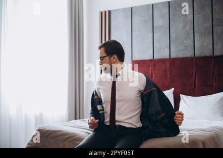 Business man with suitcase taking off jacket while sitting on bed in hotel room Stock Photo