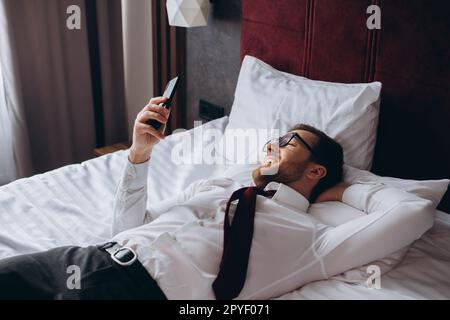 A businessman rests lying on a bed in a hotel room. A young man uses his smartphone while resting. Stock Photo