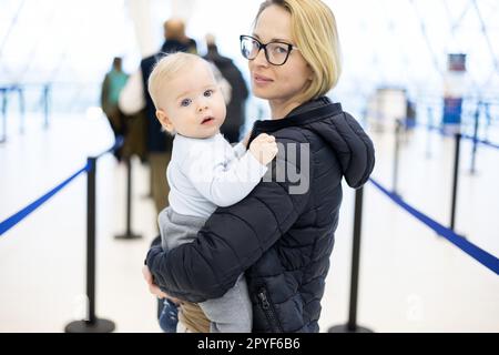 Mother carying his infant baby boy child queuing at airport terminal in passport control line at immigrations departure before moving to boarding gates to board an airplane. Travel with baby concept. Stock Photo