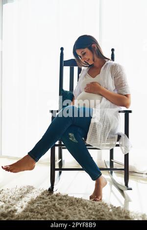 I cant wait to meet you. a happy pregnant woman cradling her belly while relaxing in a chair at home. Stock Photo