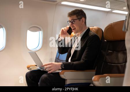Cheerful businessman in airliner using laptop computer while talking on cellphone. Stock Photo