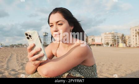 Young woman sitting on sandy beach and using mobile phone Stock Photo