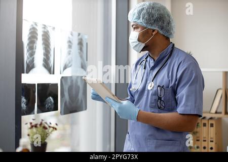 Attentive multiethnic man in medical scrubs and face mask looking at X-ray pictures while holding tablet in doctor's workplace. Physician in gloves and cap examining chest scans in hospital interior. Stock Photo