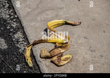 Banana peel laying on the ground - grungy grainy blacktop and sidewalk - slip and fall danger - focus on foreground. Stock Photo