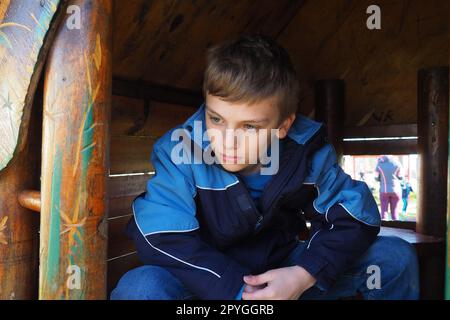 A 10-year-old Caucasian boy with blond hair and gray eyes looks out of the door of a wooden house on a children's playground. Peekaboo. The child hid. Nice face. The boy is sitting in a small house. Stock Photo